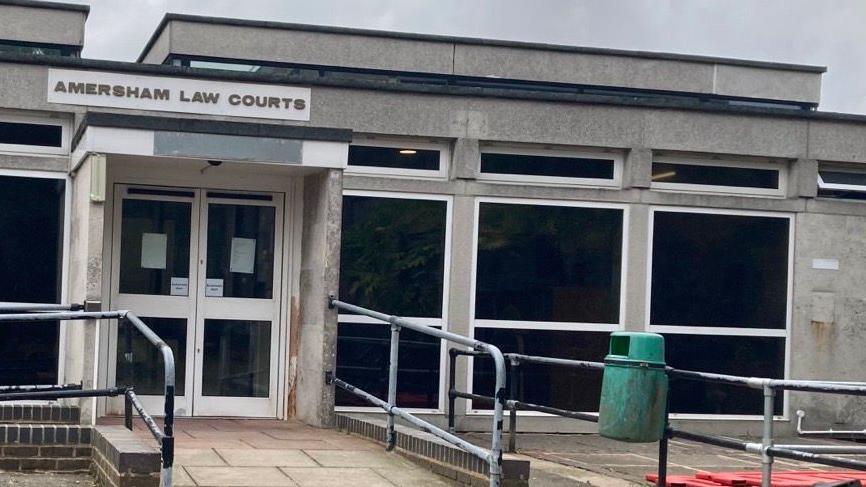 Amersham Law Courts: A grey, one-storey building. The building has white framed glass doors and white-framed windows. A grey metal fence and green waste bin stand in from of the building. A sign above the entrance says "AMERSHAM LAW COURTS",