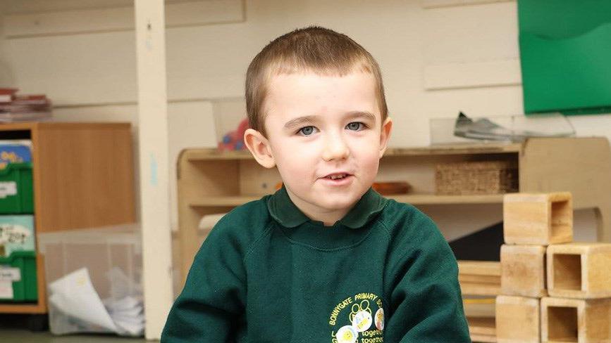Lincoln Button is wearing a green school top.  He is in a schoolroom, surrounded by various bits of bric a brac.