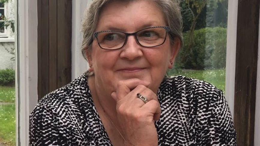 Family photo of Rachel. She is in a house with large windowns behind showing a garden. She has grey hair and glasses. Her hand is on her chin as she poses for the photo.
