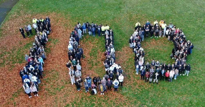 An overhead shot of a crowd of people on a piece of grass arranged to show the number 100.