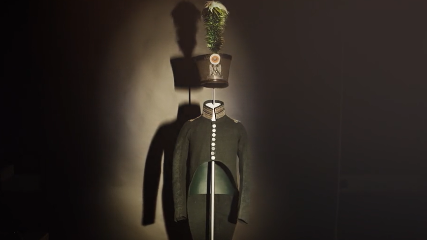 The uniform is displayed in a dark room with a single light on it. The jacket is like a wastecoat at the front, but then curves downwards into a long tail.  The hat is like a top hat, and has a large green feather on the top.