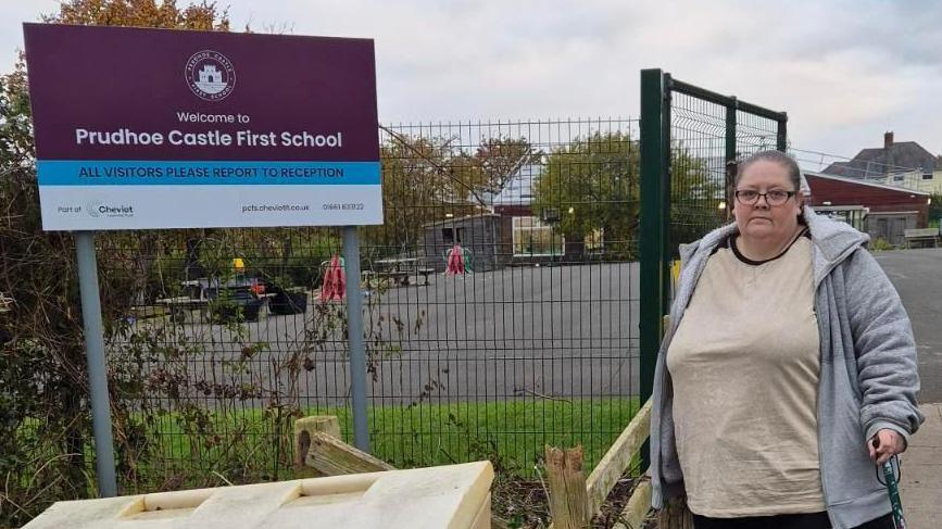 Michelle Gallon is wearing a grey hoodie and a white top. She has glasses and has hair tied back. She is holding a walking stick. She is standing by a large purple sign which says "Prudhoe Castle First School". It is daytime. School grounds can be seen in the background with tables.