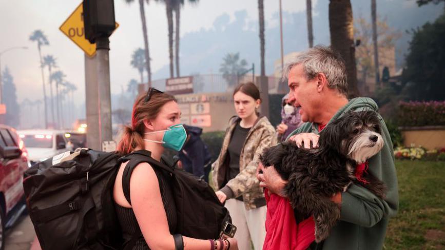 A family, two young women and an older man, stand around waiting to be evacuated ahead of wildfires in LA. The man is holding a dog, one of the women has a facemask on. They have large bags.