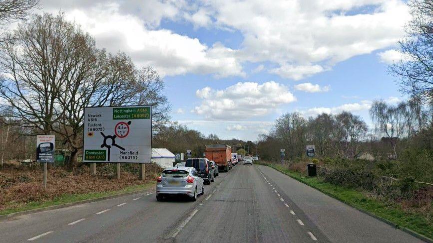 A street view of the A614 in Nottinghamshire showing a road sign for a roundabout on the left of the road. A number of cars are queuing and there are trees lining the road on both sides.