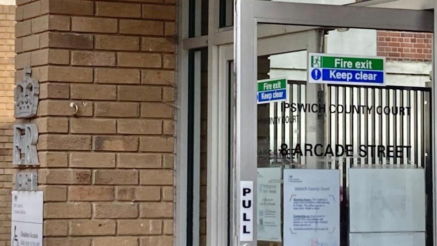 Ipswich County Court: an open grey-framed glass door with the words "IPSWICH COUNTY COURT 8 ARCADE STREET" written in black capital letters. Other signs on the door say "PULL" and "Fire exit" and "Keep clear". One the left is a brown brick wall. The silver letters "ER", an a silver sign saying "Disabled Access" are fixed to the wall