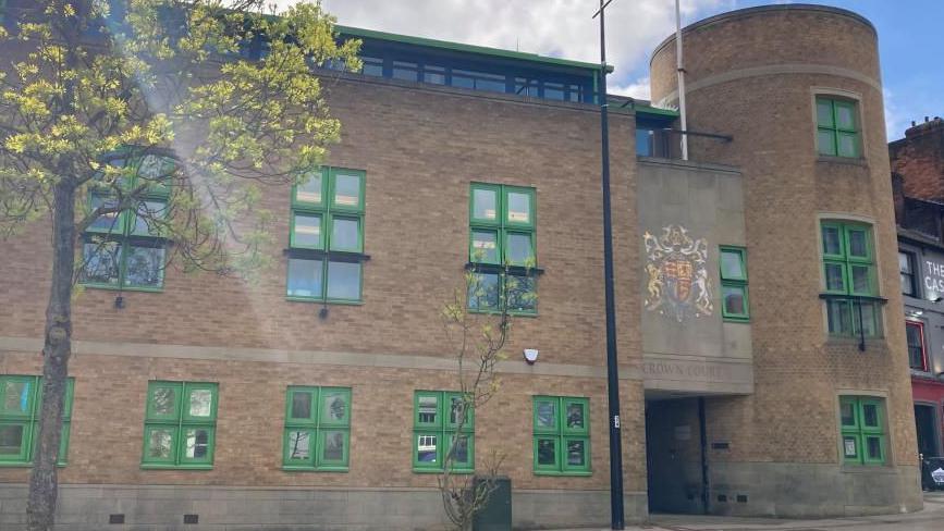Luton Crown Court: a brown-brick building with green-framed windows. The words "CROWN COURT" are written above the entrance. A tree with yellow leaves is in front of the building.