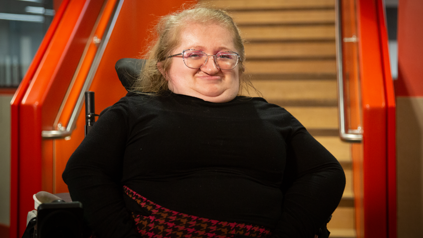 Kat Watkins in a wheelchair, smiling at the camera. She is wearing a black top with a black and red skirt in a houndstooth pattern. There are stairs behind her with the banisters painted orange. 