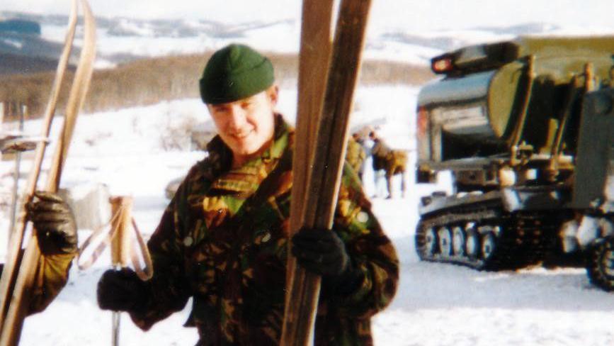 A solider wearing a camouflage jacket and green winter hat, holding skis on a snowy mountain with a military vehicle on tracks behind him.