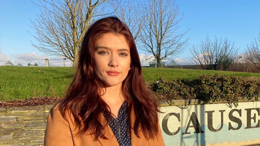 Cara Hunter looking at the camera with a neutral expression. She is wearing a brown coat. Behind her a grassy hill and a few trees.