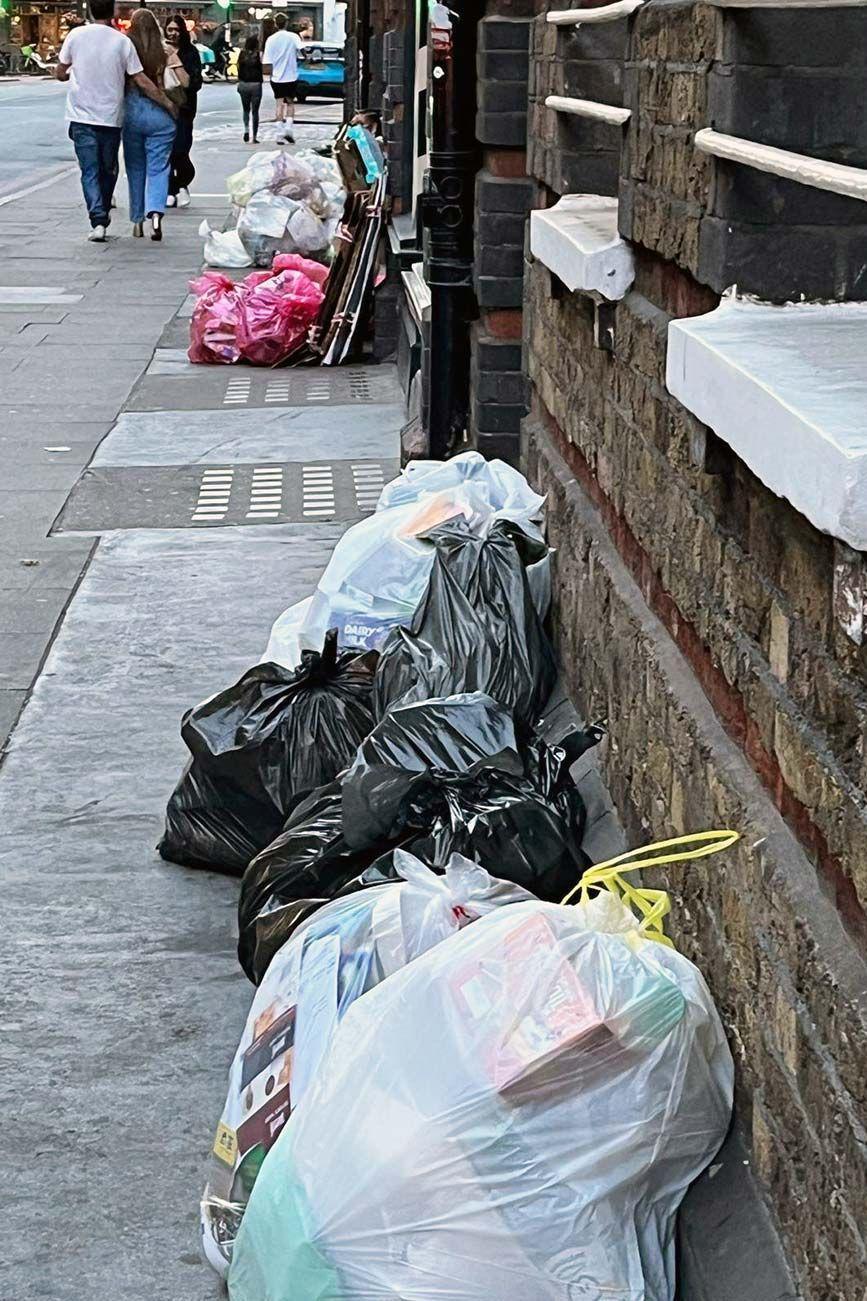 Rubbish bags lined along a pavement next to a brick building, including black and clear plastic bags filled with mixed waste. More bags, including pink ones, are visible further down the street near stacked cardboard. Several people are walking in the background.