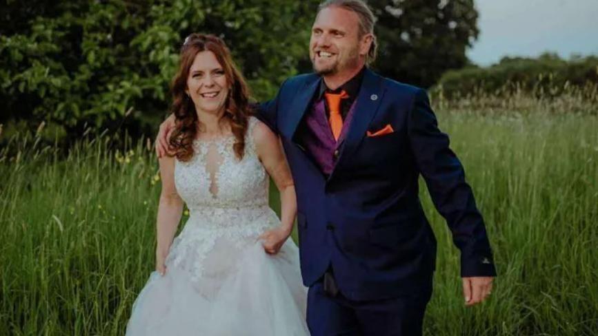 Kayleigh and Christopher Coll walking through a field of long grass and smiling - Mrs Coll is wearing a white wedding dress and Mr Coll is wearing a dark blue suit, purple waistcoat and orange tie.