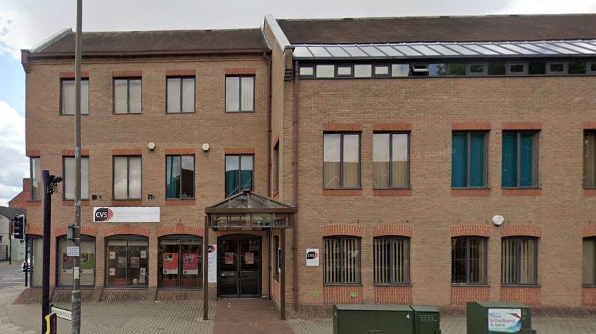 Three-storey brick building with black-framed windows equally spaced throughout. The right side of the building is set back from the left side, and there is an entrance door with a permanent gazebo-like structure in front of it stretching out into the pavement. A sign saying CVS is visible over the left-hand section of the building.