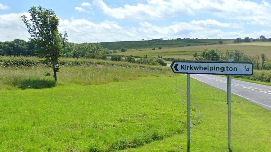 A road running through green fields. A road sign pointing to Kirkwhelpington can be seen to the right.