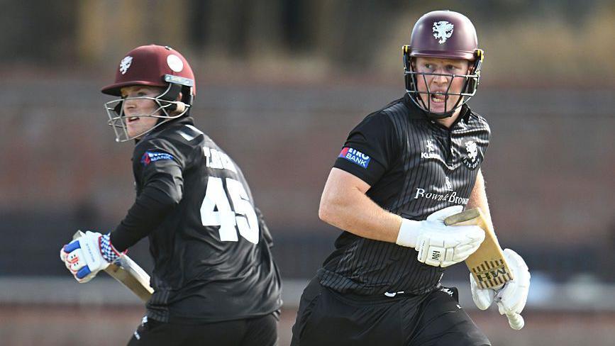 Thomas Rew (left) runs in one direction with James Rew (right) running the other as both hold bats in their hand during a match for Somerset