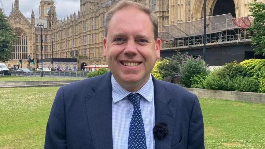 The MP stands outside the Houses of Parliament on the lawns, he wears a blue suit, shirt and tie and is smiling. He has short fair hair.