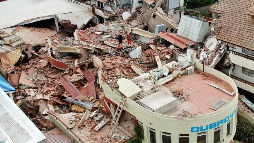 A drone image of the debris at the Dubrovnik hotel, which shows widespread damage to surrounding buildings and a lot of rubble. Rescue workers can be seen stood among debris.