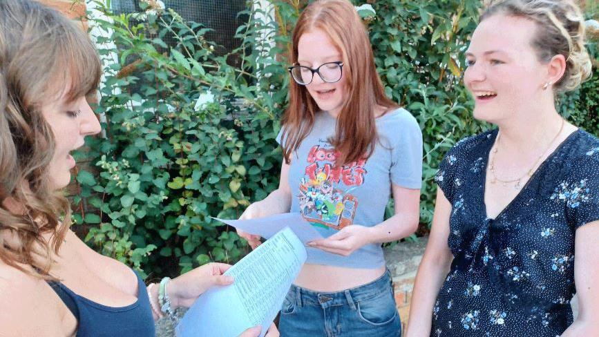 Three female students look happy as they stand together looking at their A level results on pieces of paper.