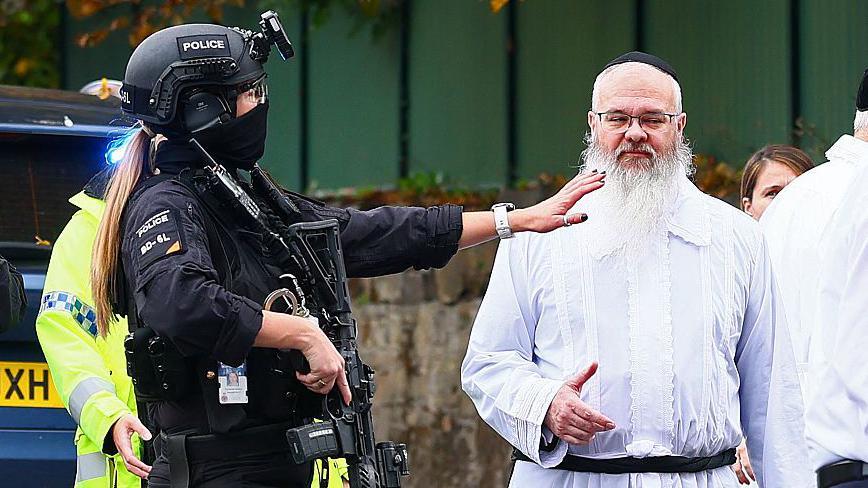 Armed police officer talk to Rabbi Walker in front of a wall with green fencing above