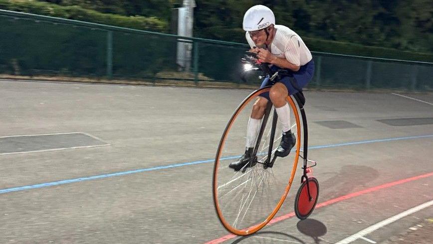 Richard Thoday on his penny farthing at Reading Velodrome. The bike is orange and he is crouched over it on the track.