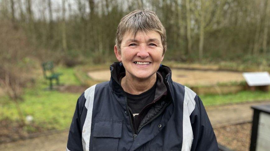 Carol Heath, wearing a black coat with grey stripes down each side, is stood with trees behind her. She has short brown hair.