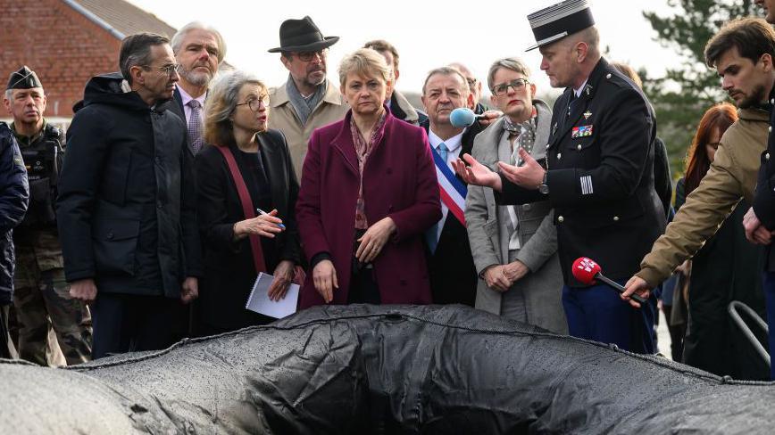 Yvette Cooper wearing a purple coat, centre, looks towards the camera at a black dingy. On the left, Bruno Retailleau looks past her to the right at a member of the army in a black coat who speaks to them both. Behind and around them are various other officials and members of the media