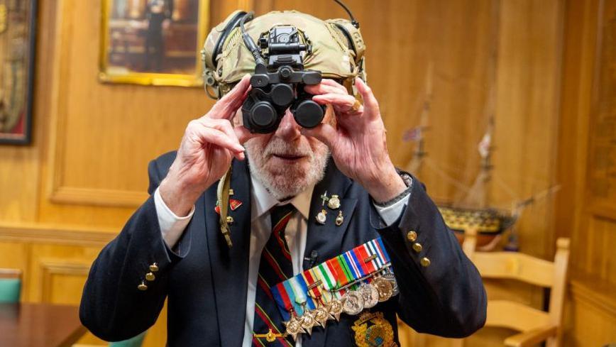 Mr Eskdale wearing a blue suit with a white shirt and navy blue and red tie. He is also wearing several medals while he wears a modern military helmet with night vision. 