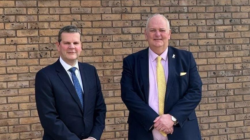 Chris Todd stands next to Jonathan Evison. Mr Todd is wearing a dark blue suit, a white shirt and a blue tie. Mr Evison is wearing a dark suit, a pink shirt and a yellow tie. They are standing in front of a brick wall.