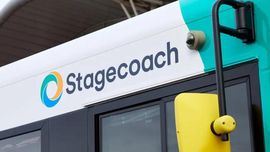 The right side of a white and mint green bus with a yellow wing mirror. The image is zoomed in above the driver's window showing the top of the window and the word Stagecoach written in black letters on the bus itself