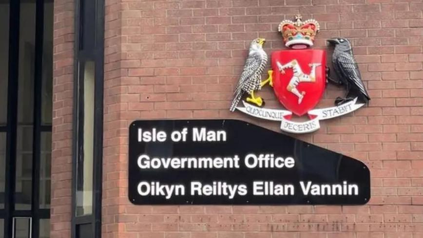 The Isle of Man Government Office sign. The wording is white on a black background and is also written in Manx. The Manx coat of arms which has a triskelion, two birds and a crown sits above the writing. It is attached to a red brick wall.