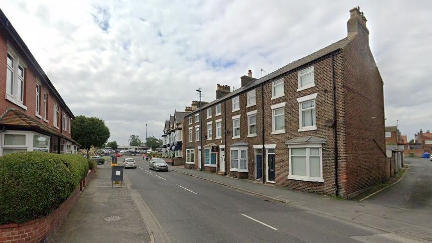 An image of a street with houses on each side. Two cars are travelling in opposite directions of the road.