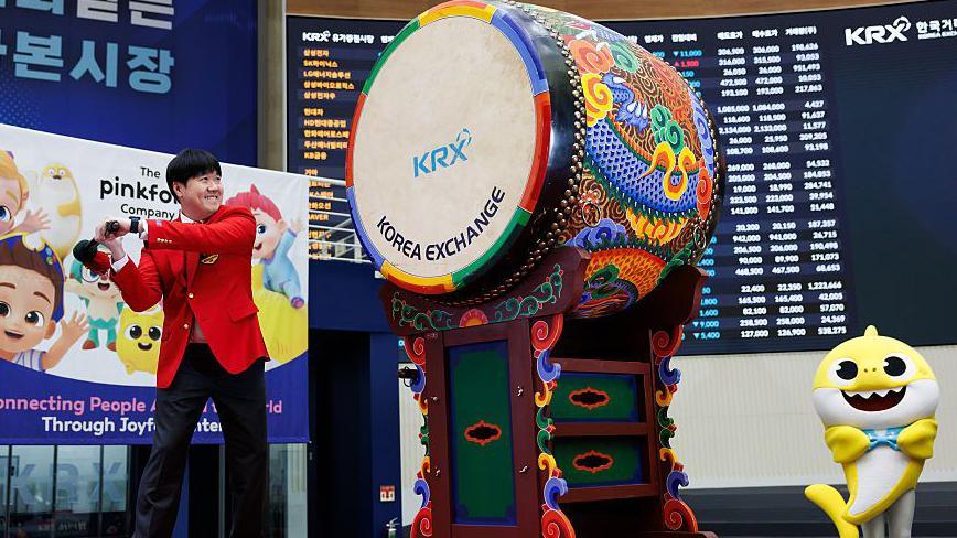 Kim Min-seok, chief executive officer of Pinkfong, strikes the drum during the firm's listing ceremony at the Korea Exchange in Seoul, South Korea, on 18 November 2025. He is dressed in a red suit as he swings a baton at a multi-coloured drum while a yellow Baby Shark mascot standing on the right of the image cheers him on.
