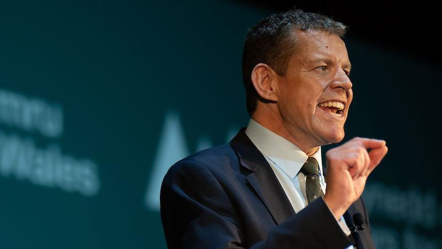 Rhun ap Iorwerth speaking. He has a dark blue suit jacket on and a light blue shirt. His right hand is up and his forefinger and thumb are together.