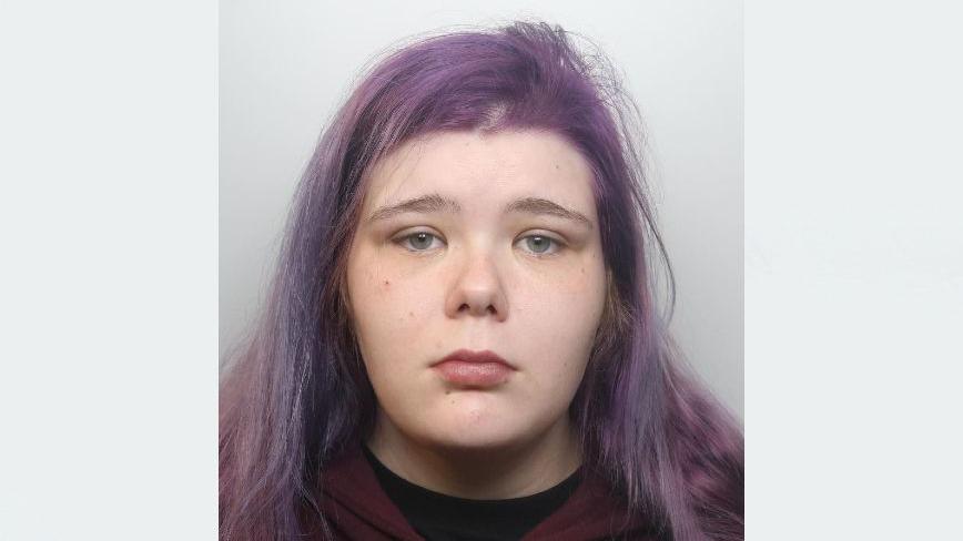 A police mugshot of Katherine Reilly, wearing a black t-shirt underneath a dark maroon hoodie. She has light blue eyes and long straight hair which is dyed purple. She is looking at the camera with a forlorn expression.