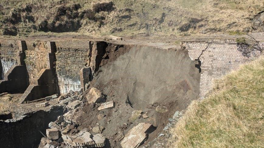 A large wall has collapsed. There is rubble and a large mudbank that has flowed out of it. The remainder of a wall is still standing on either side.