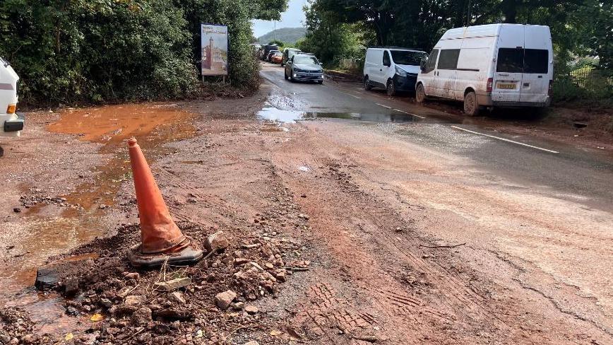 Rocks and gravel can be seen deposited in the car park along with reddish orange mud.  There is a traffic cone on the top of it. There is also a large orange puddle and a sign reading Welcome to Kingsand and Cawsand.