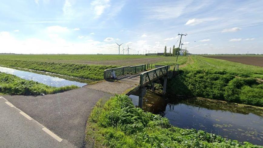 Fields span a wide landscape with a road and a bridge leading to them across a dyke, which runs parallel to a road. In the distance are wind turbines. 
