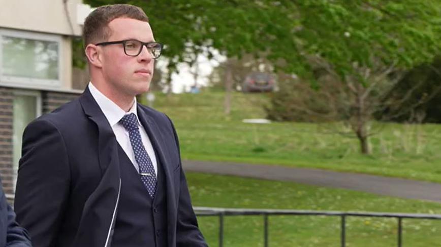 Ellis Thomas is seen walking in navy suit and tie and white shirt. He wears dark-rimmed glasses and has short brown hair.