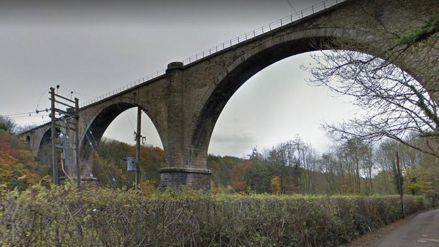 The stone Victoria Viaduct photographed from the path below. The viaduct has fencing with trees and shrubs surrounding its foundations.