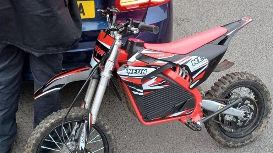 Red-and-white Neon scrambler bike seized by police resting on the boot of a navy blue car with a person wearing navy trousers and coat standing nearby.