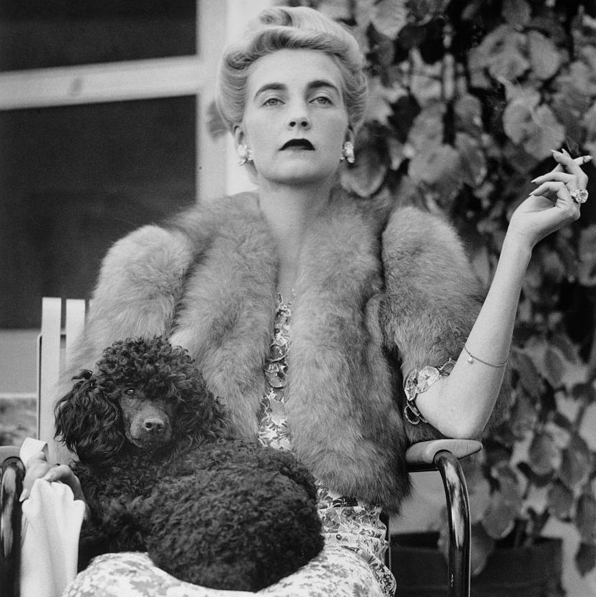 A black and white photo of a woman holding a cigarette in her left hand with a dog on her lap