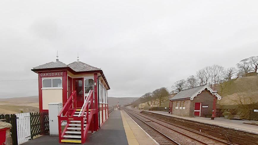 A wooden red and cream passenger waiting room stands on a platform on one side of twin railway tracks with a single storey brick building on the right hand side platform. The hills of the Yorkshire Dales can be seen in the background.