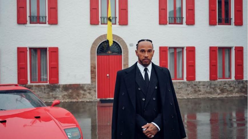 Lewis Hamilton stood alongside a Ferrari sports car while wearing a black suit and black overcoat