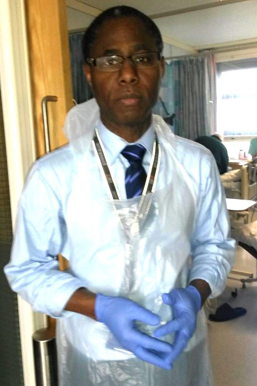 Mr Akomolafe is stnding opposite a hospital cubicle and looks straight into the camera. He is wearing glasses, a light blue shirt and dark blue striped tie, with a plastic apron on top. He has disposable blue gloves on his hands.