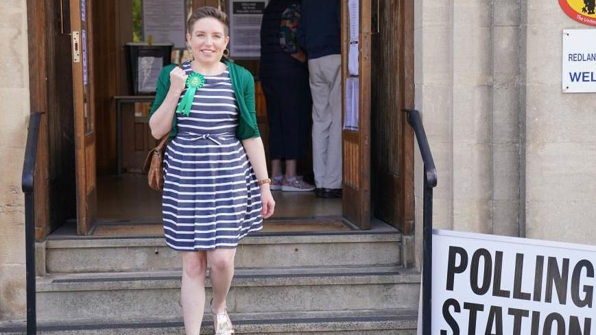 Carla Denyer smiling while wearing in her green rosette outside a polling station