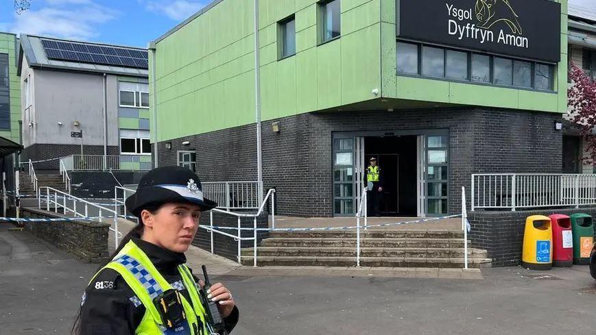 Ysgol Dyffryn Aman with a police officer holding a walkie talkie, standing in front of police tape that's draped on the railings in front of the main entrance. A police officer can also be see in the doorway of the entrance
