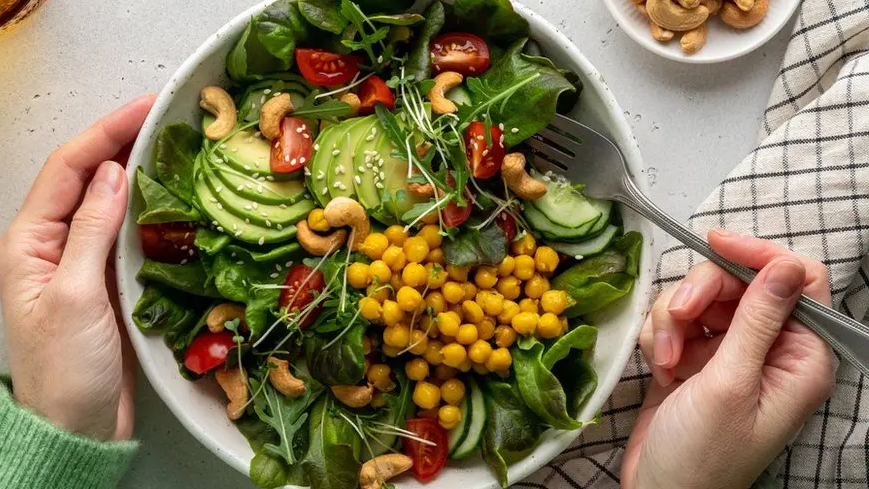 An above view of a white bowl which has a salad in, containing cucumber, avocado, tomatoes and leaves. A person is holding a fork with their right hand.
