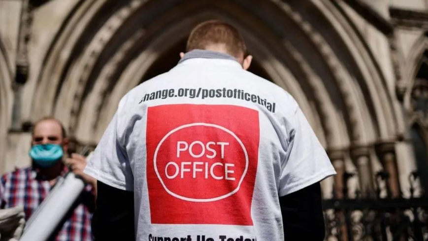 A campaigner outside court from behind, wearing a t-shirt emblazoned with a Post Office logo and slogan