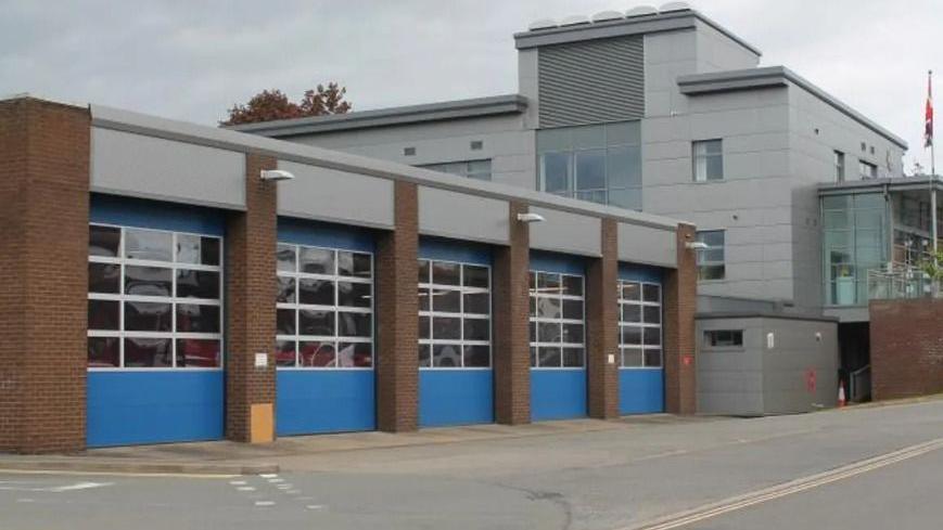 A fire station pictured from the road outside. A high appliance bay can be seen, with four large doors that are blue in between brick pillars. A large grey building is on the right.