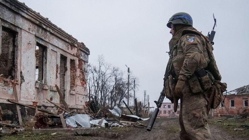 Lone Ukrainian in military fatigues stands outdoors as he looks at destroyed building. More damaged buildings are visible in the background