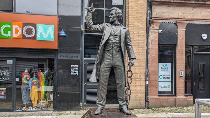 A life-size statue of Frederick Douglass in Belfast which was unveiled in July 2023.  The bronze sculpture shows a tall, well-dressed black man holding a chain in one hand and raising his other hand, as if gesticulating during a speech. 